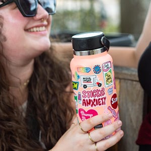 A woman with curly hair is holding a pink water bottle with various stickers on it, smiling and looking at the camera.