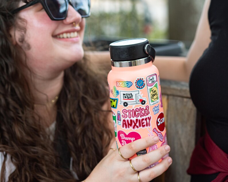 A woman with curly hair is holding a pink water bottle with various stickers on it, smiling and looking at the camera.