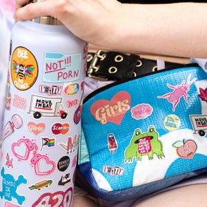 a person holding a water bottle with various stickers on it, and a bag with stickers on it. The person is wearing a black belt.