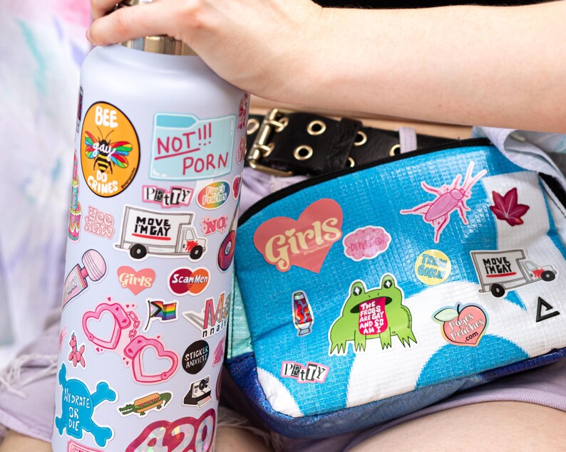 a person holding a water bottle with various stickers on it, and a bag with stickers on it. The person is wearing a black belt.