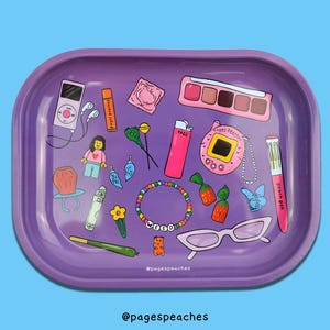 May include: A rectangular purple rolling tray featuring a colorful illustration of various items including a digital music player, a pink lighter, a beaded bracelet with the word WEED, a multi-colored pen, eyeglasses, and a small toy figure.
