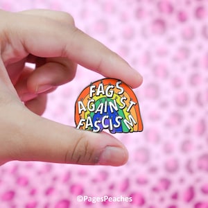 May include: A rainbow-shaped enamel pin with the text "Fags Against Fascism" in white. The pin features a rainbow design with colors including red, orange, yellow, green, blue, and purple. The pin is held against a pink background.