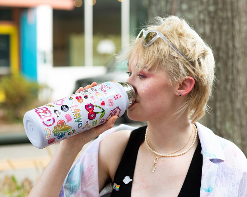 A woman with short blonde hair is drinking from a colorful water bottle with various stickers on it.