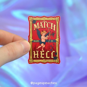 Peut inclure: Autocollant rectangulaire avec un fond rouge et une bordure dorée. L'autocollant présente le texte "MATCH MADE IN HELL" en lettres dorées. Une illustration d'un chérubin avec des ailes et un arc et des flèches est également présente.