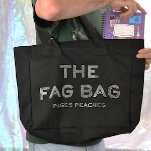 Op de afbeelding: Een zwarte canvas tas met de tekst THE FAG BAG en PAGES PEACHES in zilverkleurige strass-steentjes. De tas heeft twee zwarte stoffen hengsels en bevat een paars spiraalnotitieboek met diverse kleurrijke grafische illustraties.