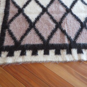May include: A textured rug with a geometric diamond pattern. The design features black, white, and mauve diamonds. The rug has a fringed edge and is made of a soft, fuzzy material.