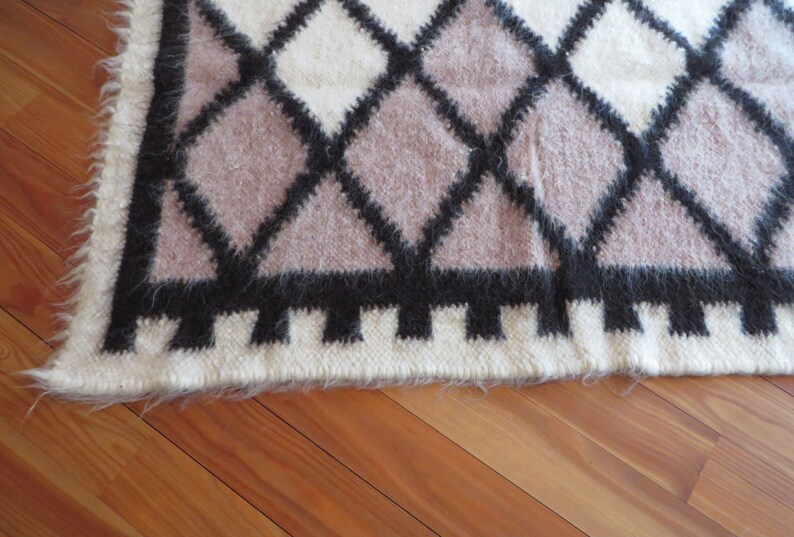 May include: A textured rug with a geometric diamond pattern. The design features black, white, and mauve diamonds. The rug has a fringed edge and is made of a soft, fuzzy material.