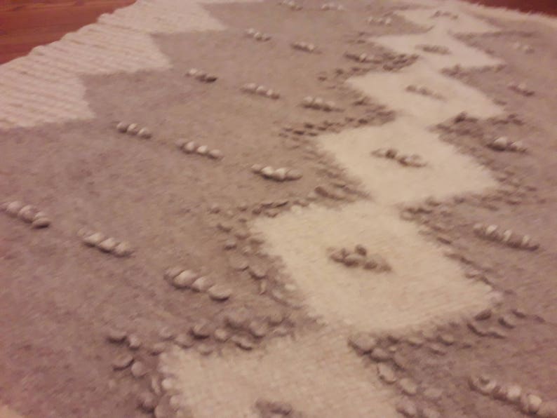 May include: A close-up of a beige and gray rug with a textured, woven design. The rug has a pattern of diagonal lines and small, raised bumps.