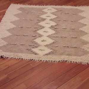 May include: A white and gray woven rug with a geometric pattern. The rug has a fringe border.