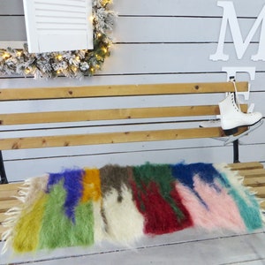 May include: A wooden bench with a colorful, fluffy rug on top. The rug is made of different colored strips of fur, including green, yellow, red, blue, and pink. A pair of white ice skates is hanging on the back of the bench.