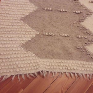 May include: A white and gray woven rug with a geometric pattern. The rug has a fringe border and is made of a soft, textured material.