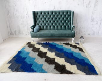 May include: A teal button-back sofa with a high back and silver legs. A blue, grey, and black patterned rug is on the floor. A small blue cushion rests on the sofa. The rug has a geometric design.