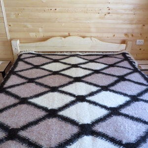 May include: A bed with a light-colored wooden headboard and matching nightstands. The bed is covered with a blanket featuring a diamond pattern in shades of white, gray, and black. The room has light wood paneling.