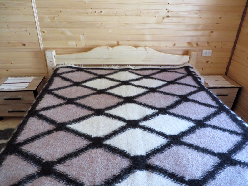May include: A bed with a light-colored wooden headboard and matching nightstands. The bed is covered with a blanket featuring a diamond pattern in shades of white, gray, and black. The room has light wood paneling.