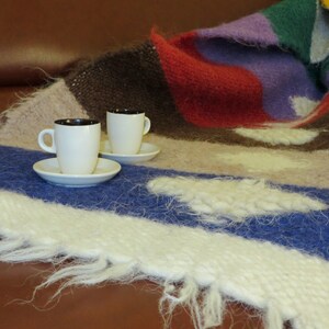 May include: Two white ceramic coffee cups with black rims sit on a white saucer on a multi-colored knitted blanket. The blanket has shades of brown, red, purple, and blue.