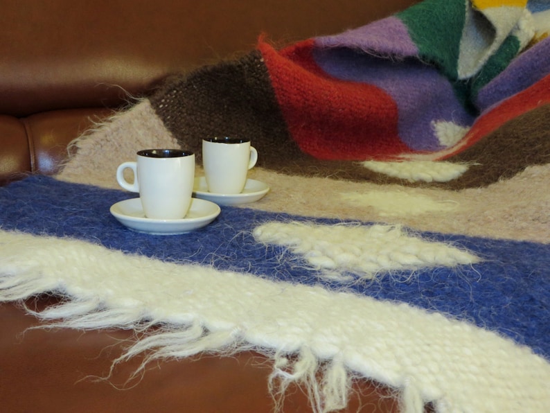May include: Two white ceramic coffee cups with black rims sit on a white saucer on a multi-colored knitted blanket. The blanket has shades of brown, red, purple, and blue.