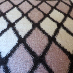May include: Close-up of a soft, textured blanket with a repeating diamond pattern. The diamonds are in shades of white, light pink, and beige, outlined with dark brown stitching. The blanket has a cozy, handmade appearance.