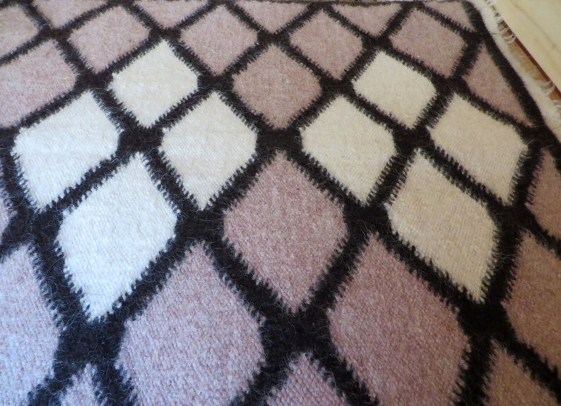 May include: Close-up of a soft, textured blanket with a repeating diamond pattern. The diamonds are in shades of white, light pink, and beige, outlined with dark brown stitching. The blanket has a cozy, handmade appearance.