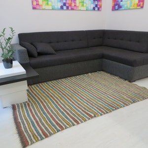 Puede incluir: Una alfombra rayada en tonos de marr&oacute;n, amarillo, azul y blanco se coloca delante de un sof&aacute; de esquina gris. Una peque&ntilde;a mesa blanca con una tapa negra se encuentra a la izquierda del sof&aacute;.