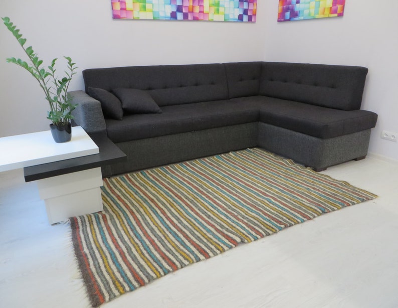 Puede incluir: Una alfombra rayada en tonos de marr&oacute;n, amarillo, azul y blanco se coloca delante de un sof&aacute; de esquina gris. Una peque&ntilde;a mesa blanca con una tapa negra se encuentra a la izquierda del sof&aacute;.