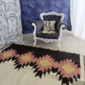 Puede incluir: Una alfombra de patrones blancos y marrones con acentos rosados y amarillos. Un sillón gris con marco blanco y una almohada con la palabra "HOME" en ella se encuentra frente a la alfombra.