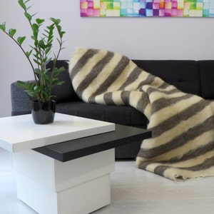 Op de afbeelding: Een wit en grijs gestreept wollen deken gedrapeerd over een zwarte bank. Het deken is gemaakt van wol en heeft een zachte, pluche textuur. Een kleine witte en zwarte salontafel staat op de voorgrond.