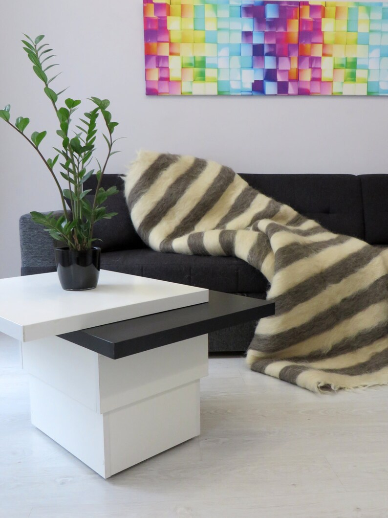 Op de afbeelding: Een wit en grijs gestreept wollen deken gedrapeerd over een zwarte bank. Het deken is gemaakt van wol en heeft een zachte, pluche textuur. Een kleine witte en zwarte salontafel staat op de voorgrond.