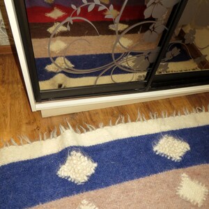 May include: A close-up of a blue, brown, and white striped rug with white, fluffy, diamond-shaped designs. The rug is made of wool and has a fringed edge.