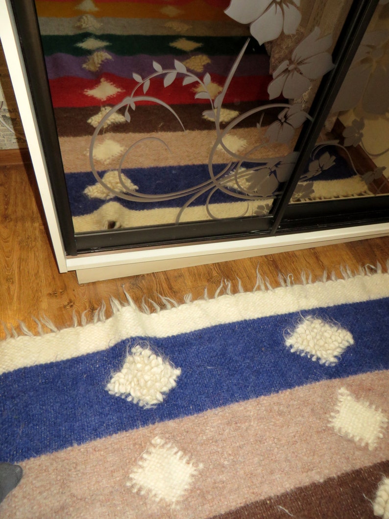 May include: A close-up of a blue, brown, and white striped rug with white, fluffy, diamond-shaped designs. The rug is made of wool and has a fringed edge.