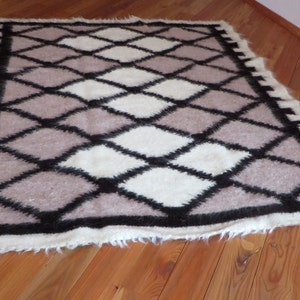 May include: A rectangular rug with a geometric diamond pattern. The rug features a combination of white, light brown, and black diamonds. The border is white with black accents. The rug is made of a soft, textured material.