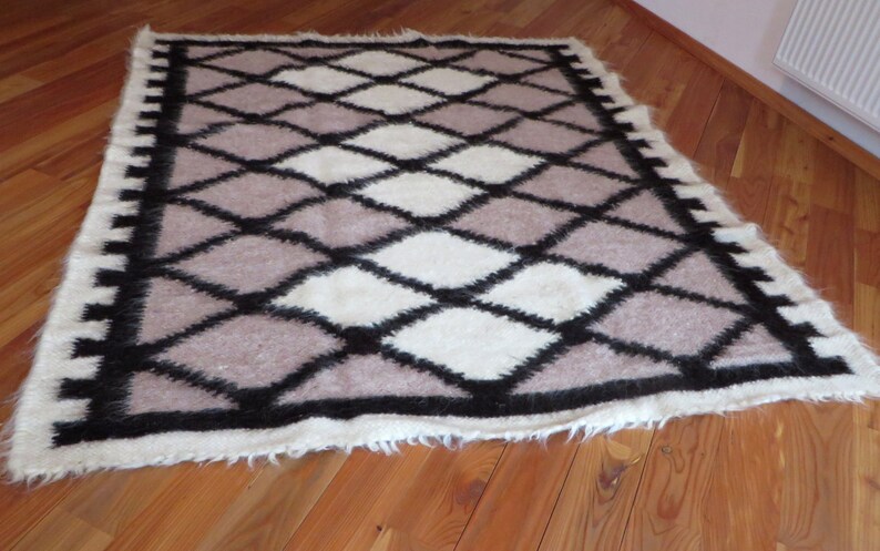 May include: A rectangular rug with a geometric diamond pattern. The rug features a combination of white, light brown, and black diamonds. The border is white with black accents. The rug is made of a soft, textured material.