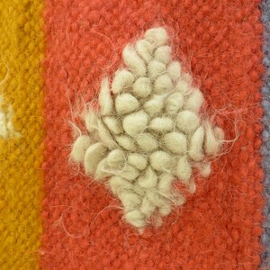 May include: Close-up of a red, yellow, and blue woven rug with a white, fluffy, diamond-shaped design in the center.