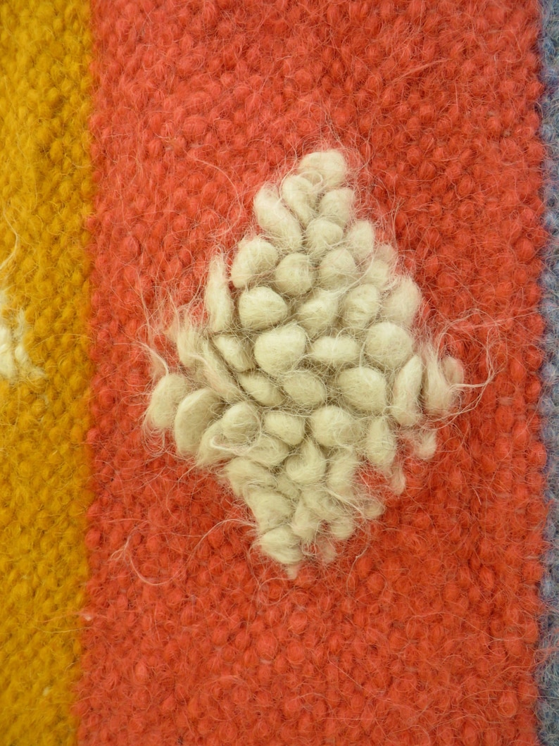 May include: Close-up of a red, yellow, and blue woven rug with a white, fluffy, diamond-shaped design in the center.