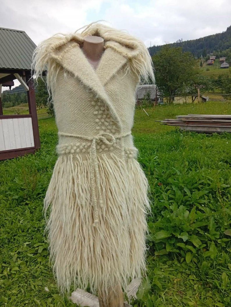 K&ouml;nnte beinhalten: Cremefarbene, &auml;rmellose Weste aus Wolle mit Schalkragen und Fransenrock. Die Weste hat eine gewebte Textur mit einem G&uuml;rtel in der Taille. Das Kleidungsst&uuml;ck wird auf einer Schaufensterpuppe in einer grasbewachsenen Umgebung im Freien pr&auml;sentiert.