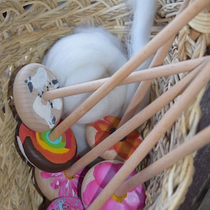 May include: A wicker basket filled with colorful wooden spinning wheels and white wool roving. The spinning wheels have painted designs, including a rainbow, a flower, and a peacock.