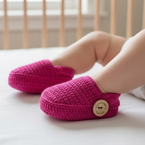 May include: A pair of bright pink crochet baby booties. The booties are made of a textured yarn and feature a wooden button on the side. The baby is lying on a white surface.