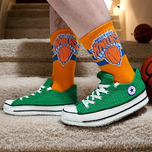 May include: Green crocheted Converse sneakers with white laces and a white sole. The sneakers are worn with orange socks that have the New York Knicks logo on them.