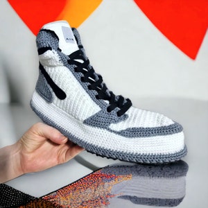 May include: A white and gray crocheted high-top sneaker with black laces and a black Nike swoosh logo. The shoe has the word "AIR" printed on the side.
