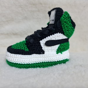 Op de afbeelding: Gehaakte babyschoen in groen, wit en zwart met het woord "AIR" aan de zijkant.
