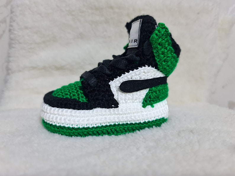 Op de afbeelding: Gehaakte babyschoen in groen, wit en zwart met het woord "AIR" aan de zijkant.