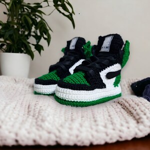 Op de afbeelding: Groene, zwarte en witte gehaakte babyschoentjes met het woord "AIR" aan de zijkant.