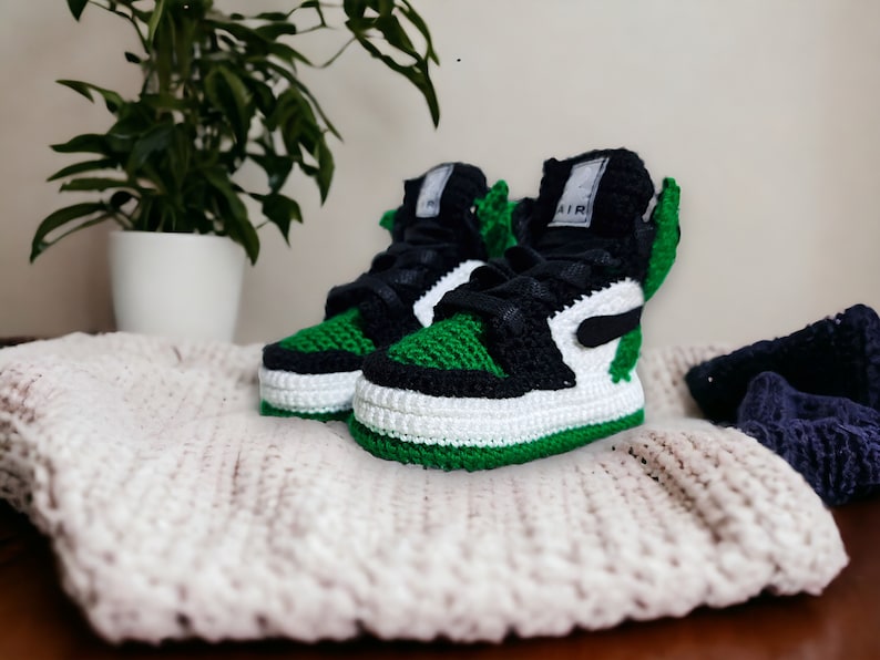 Op de afbeelding: Groene, zwarte en witte gehaakte babyschoentjes met het woord "AIR" aan de zijkant.