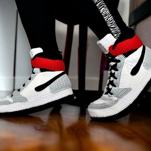 May include: White, gray, and black crocheted high-top sneakers with red accents. The sneakers are made to look like a popular athletic shoe brand.
