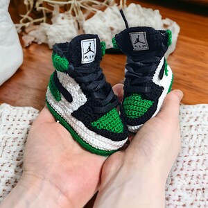Op de afbeelding: Een paar groene, witte en zwarte gehaakte babyschoentjes met het woord "AIR" aan de zijkant.