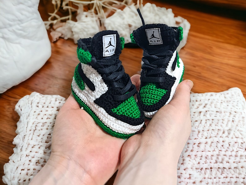 Op de afbeelding: Een paar groene, witte en zwarte gehaakte babyschoentjes met het woord "AIR" aan de zijkant.