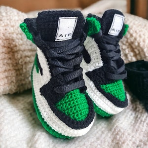 Op de afbeelding: Een paar groene, witte en zwarte gehaakte babyschoentjes met het woord "AIR" aan de zijkant.
