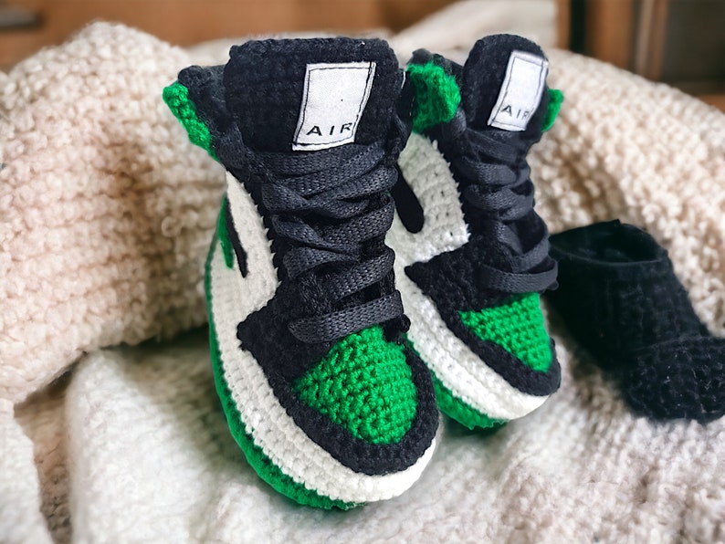 Op de afbeelding: Een paar groene, witte en zwarte gehaakte babyschoentjes met het woord "AIR" aan de zijkant.
