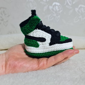 Op de afbeelding: Een groene, witte en zwarte gehaakte sneaker. De sneaker is klein en wordt in een hand gehouden.