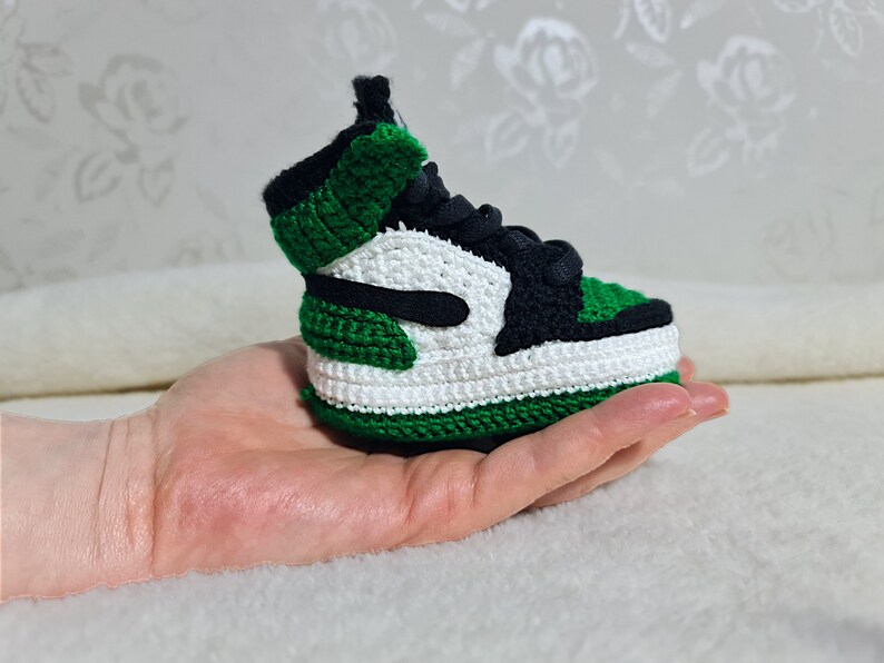 Op de afbeelding: Een groene, witte en zwarte gehaakte sneaker. De sneaker is klein en wordt in een hand gehouden.