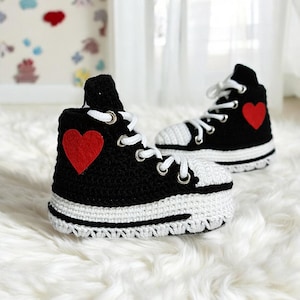 May include: A pair of black and white crocheted baby shoes with red heart accents. The shoes are high-top style and have a white sole.